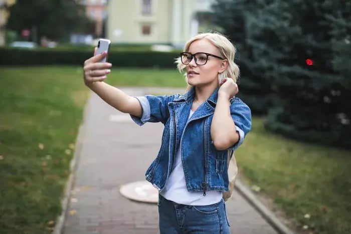 Blonde woman testing virtual eyeglass frames on her phone while walking outdoors.